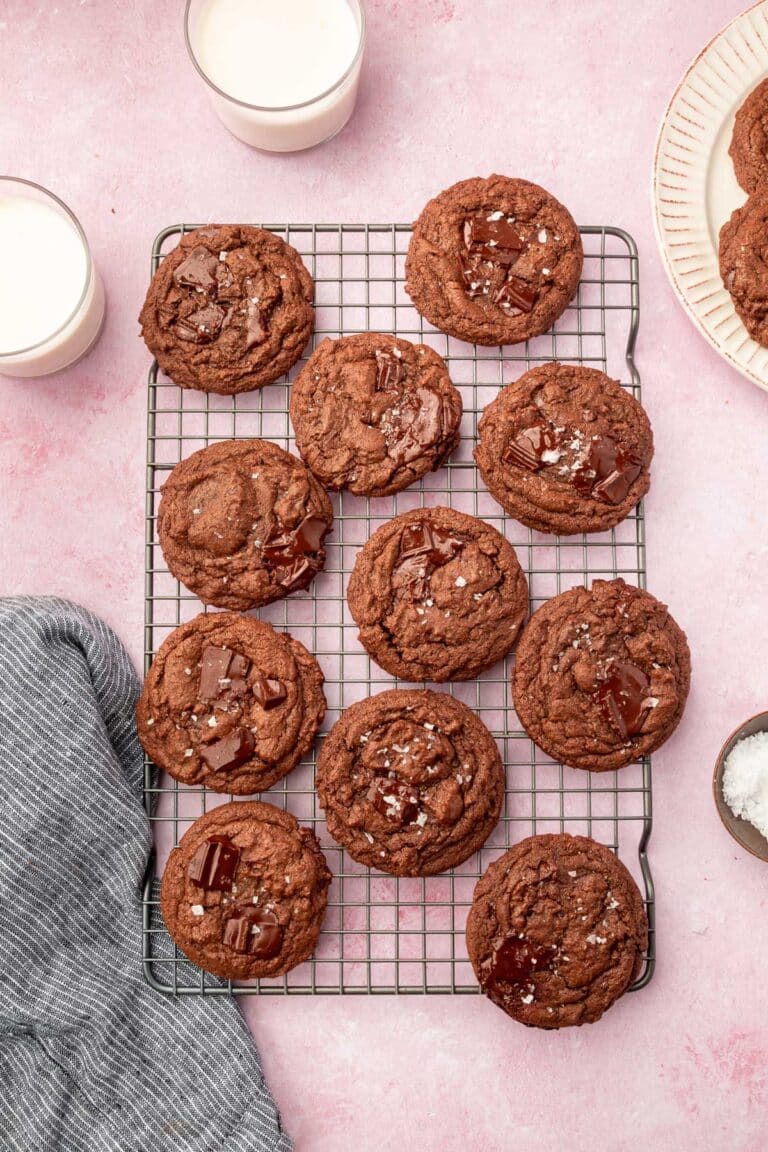 A cooling rack on a pink table filled with gluten-free chocolate chunk cookies topped with flaky sea salt with glasses of milk on the side.