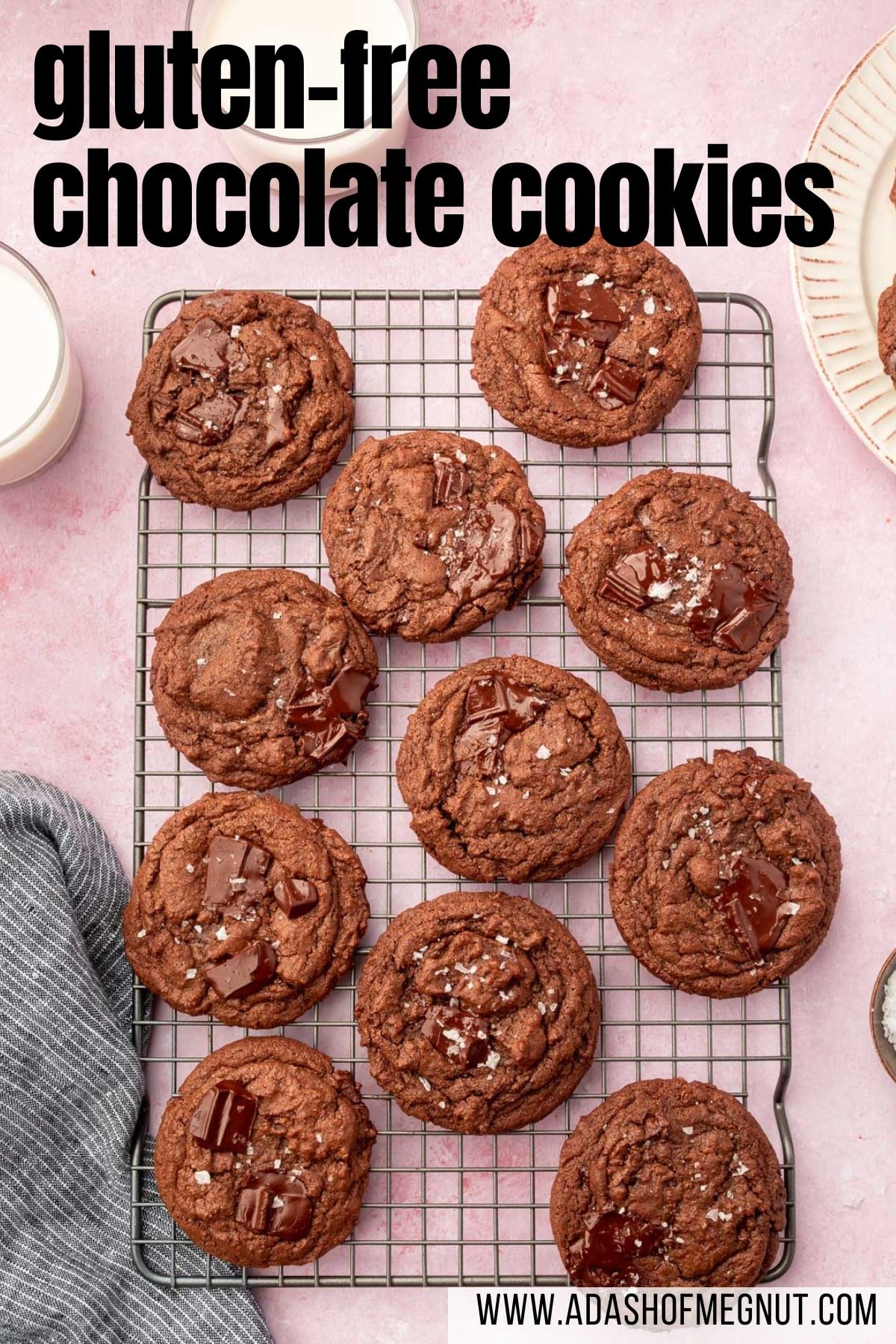 A cooling rack on a pink table filled with gluten-free chocolate chunk cookies topped with flaky sea salt with glasses of milk on the side.