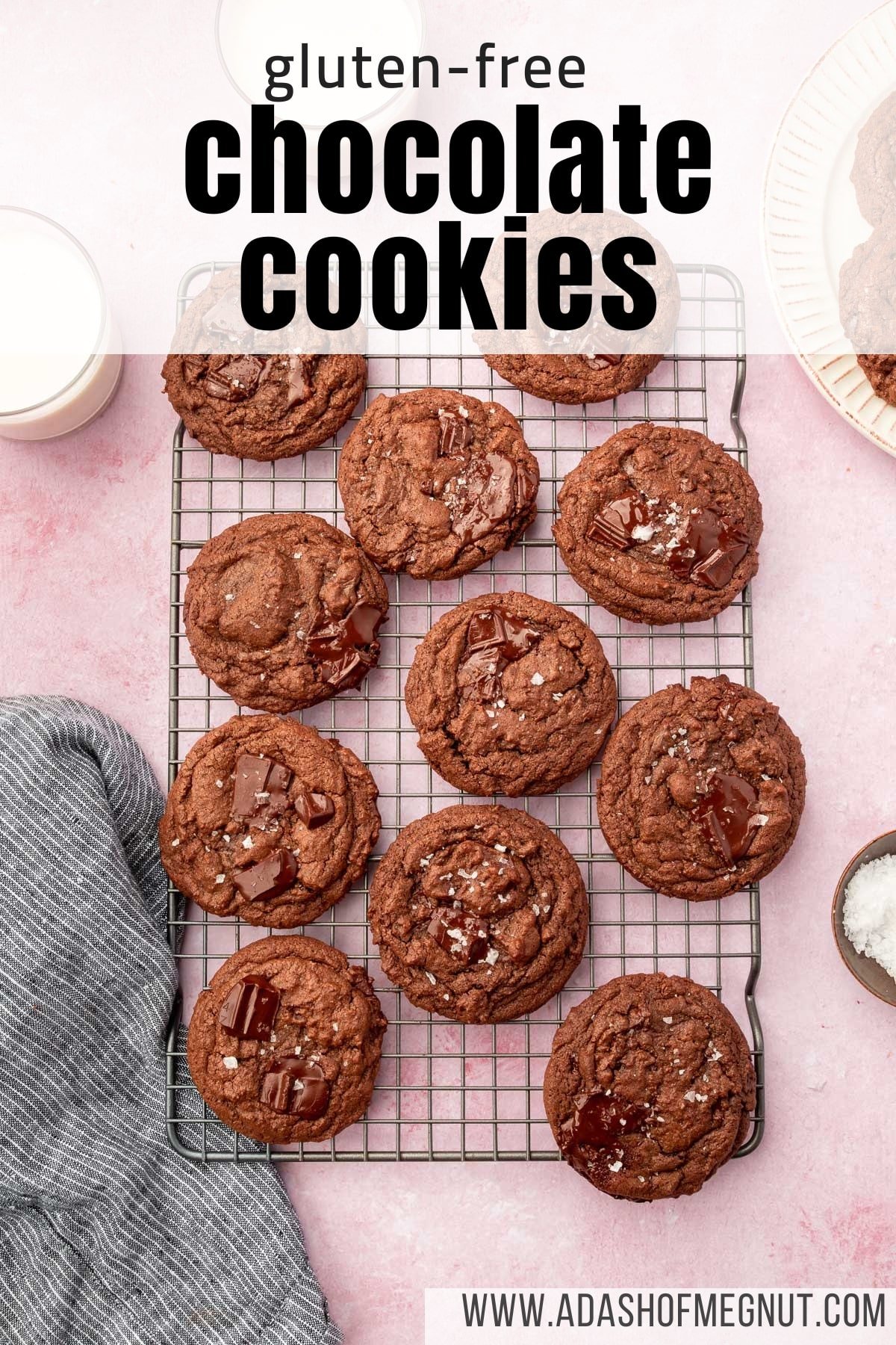 A cooling rack on a pink table filled with gluten-free chocolate chunk cookies topped with flaky sea salt with glasses of milk on the side.