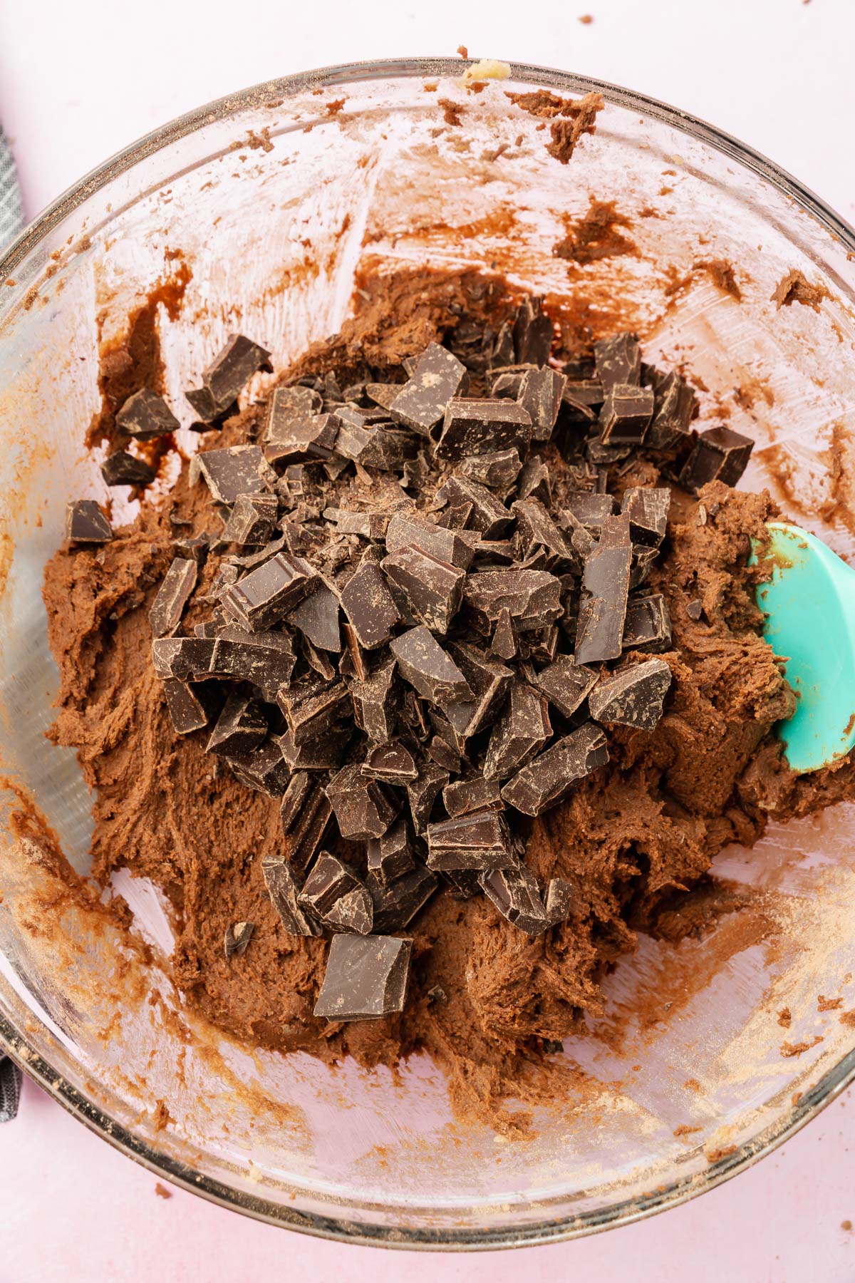 A gluten-free chocolate cookie dough topped with chopped chocolate in a glass mixing bowl with a blue spatula.