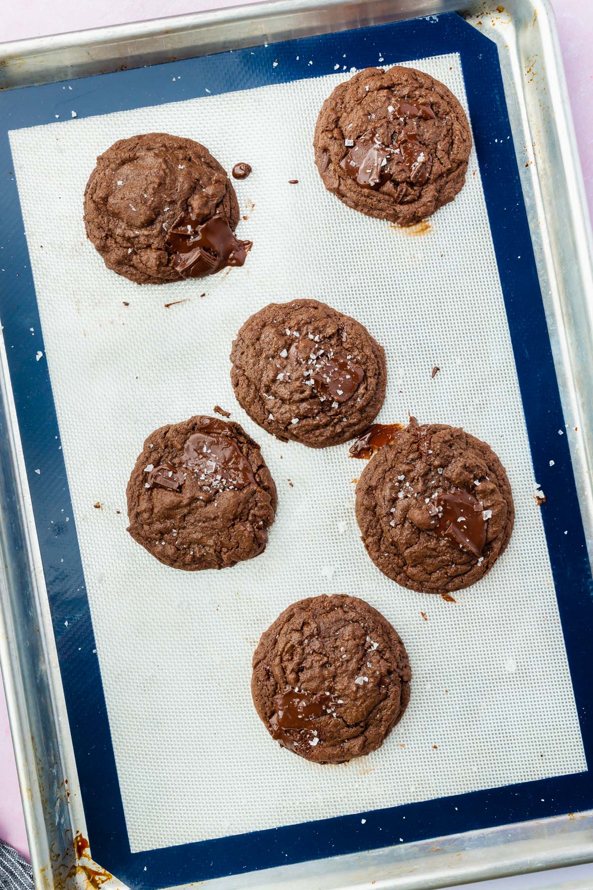 A silpat lined baking sheet topped with 6 gluten-free chocolate cookies topped with flaky sea salt.