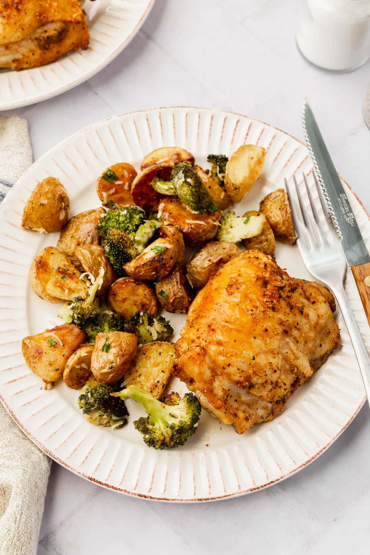 A dinner plate with a single crispy chicken thigh, a side of roasted baby potatoes and broccoli, and a fork and knife.