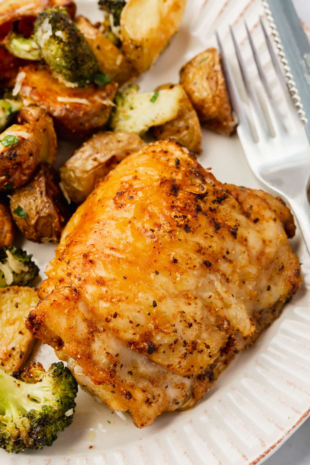 A closeup of a crispy air fryer chicken thigh on a plate with roasted potatoes and broccoli.