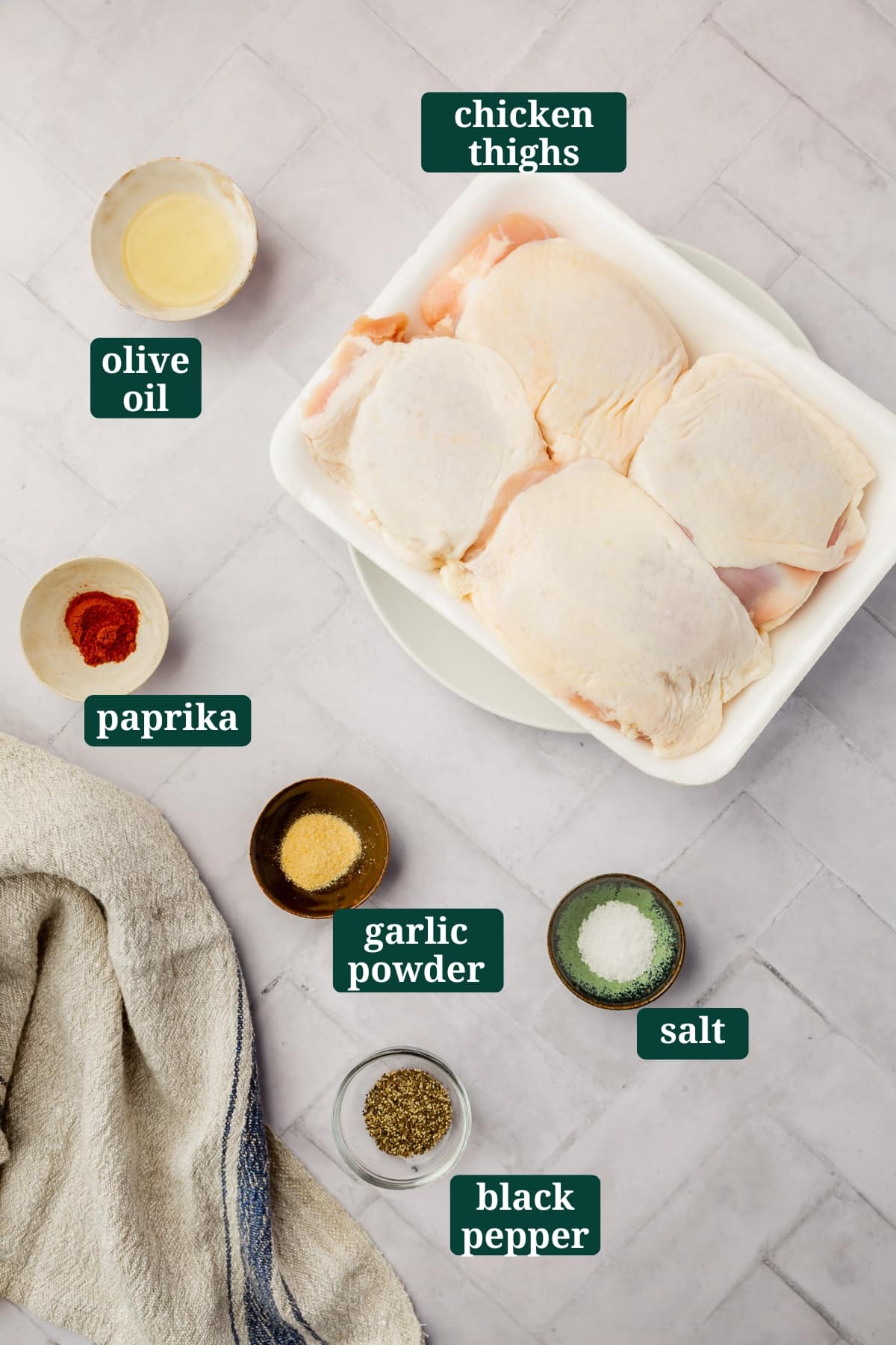 Ingredients in small bowls to make air fryer crispy chicken thighs including bone-in skin-on chicken thighs, olive oil, paprika, garlic powder, salt and black pepper with text overlays over each ingredient.
