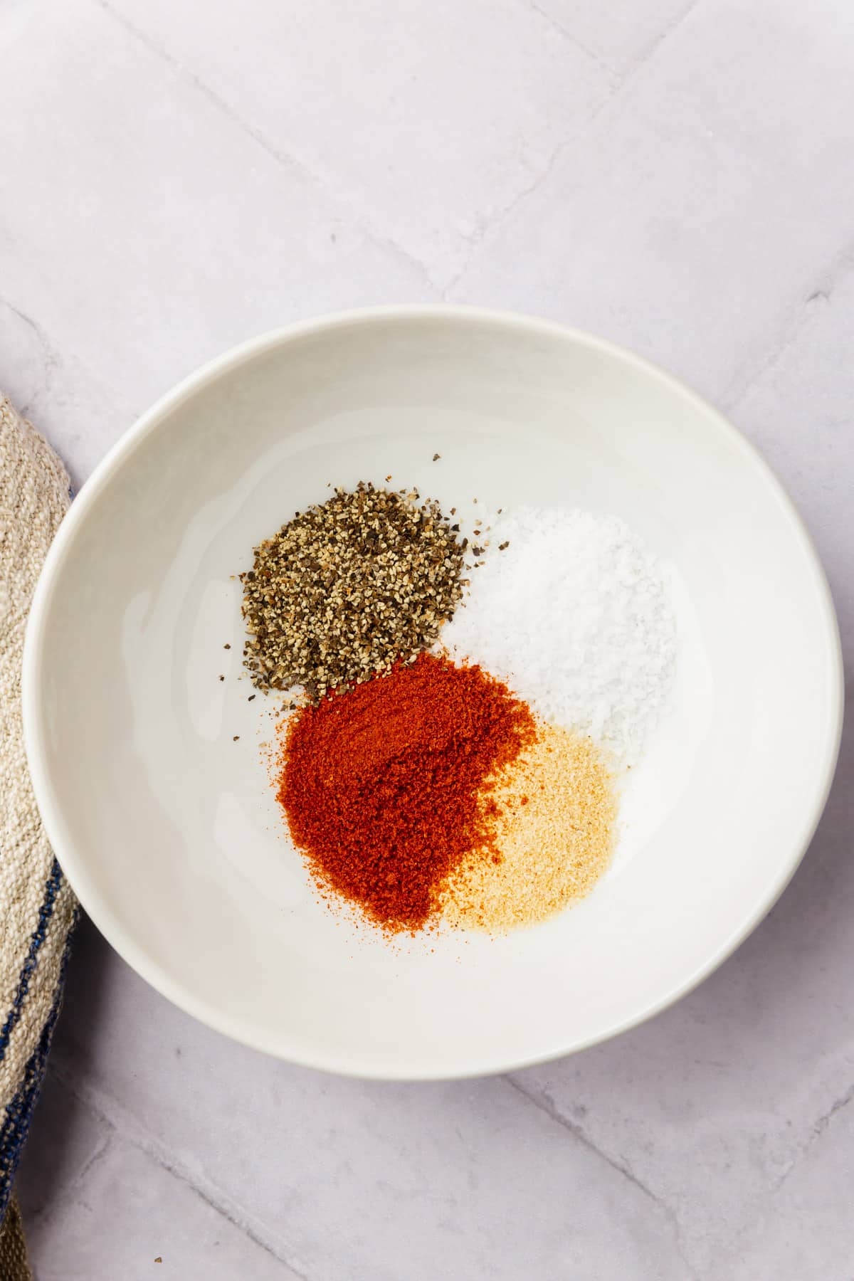 Black pepper, salt, paprika, and garlic powder in a white ceramic bowl before mixing together.