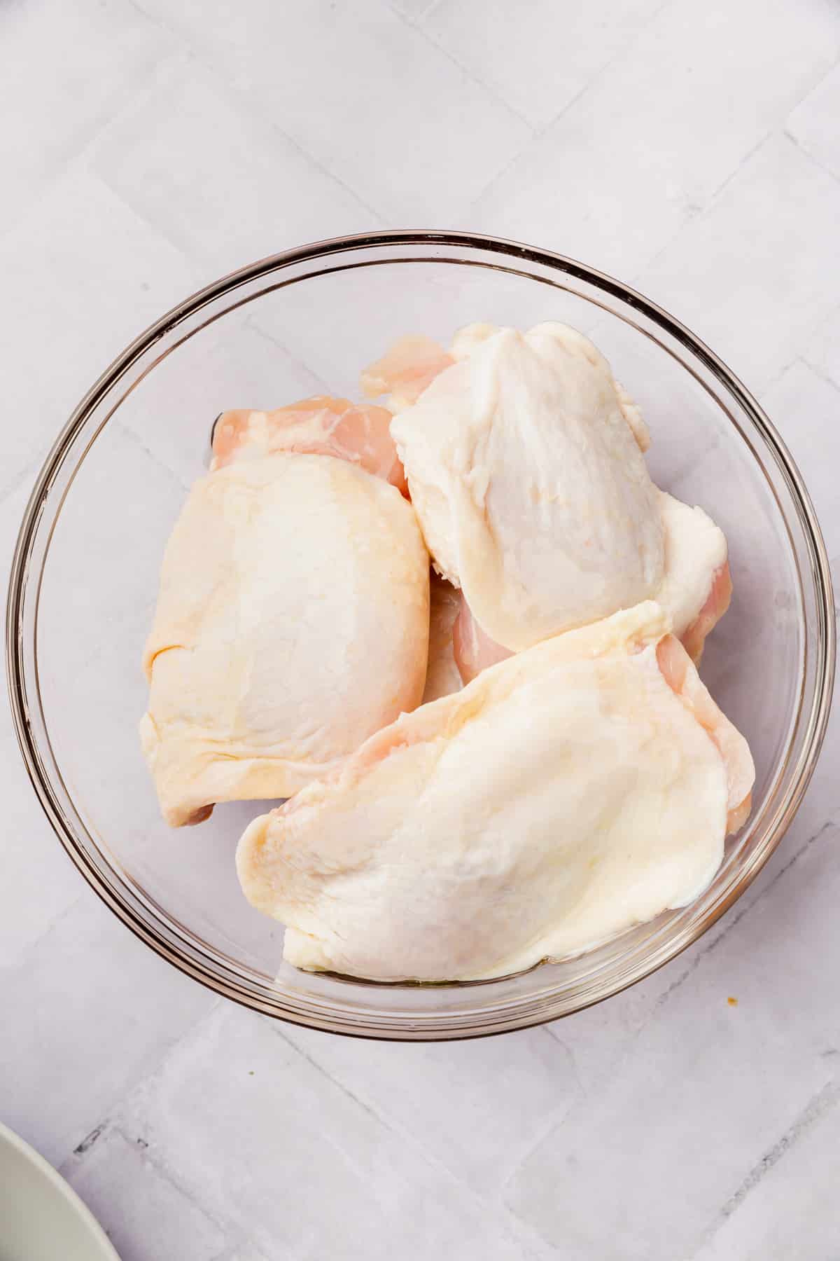 Raw chicken thighs topped with olive oil in a glass mixing bowl.