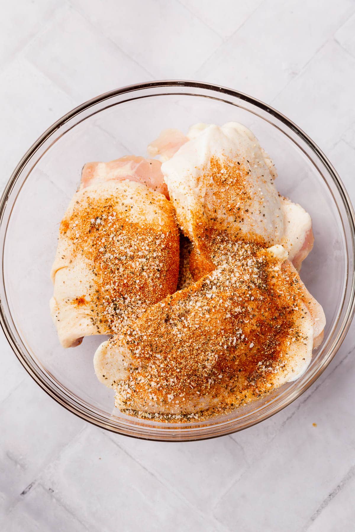 Raw chicken thighs topped with a seasoning blend before mixing together in a glass mixing bowl.