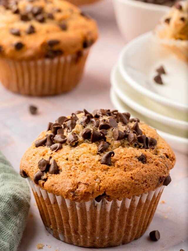 A single gluten-free mini chocolate chip muffin with a stack of dessert plates behind it.