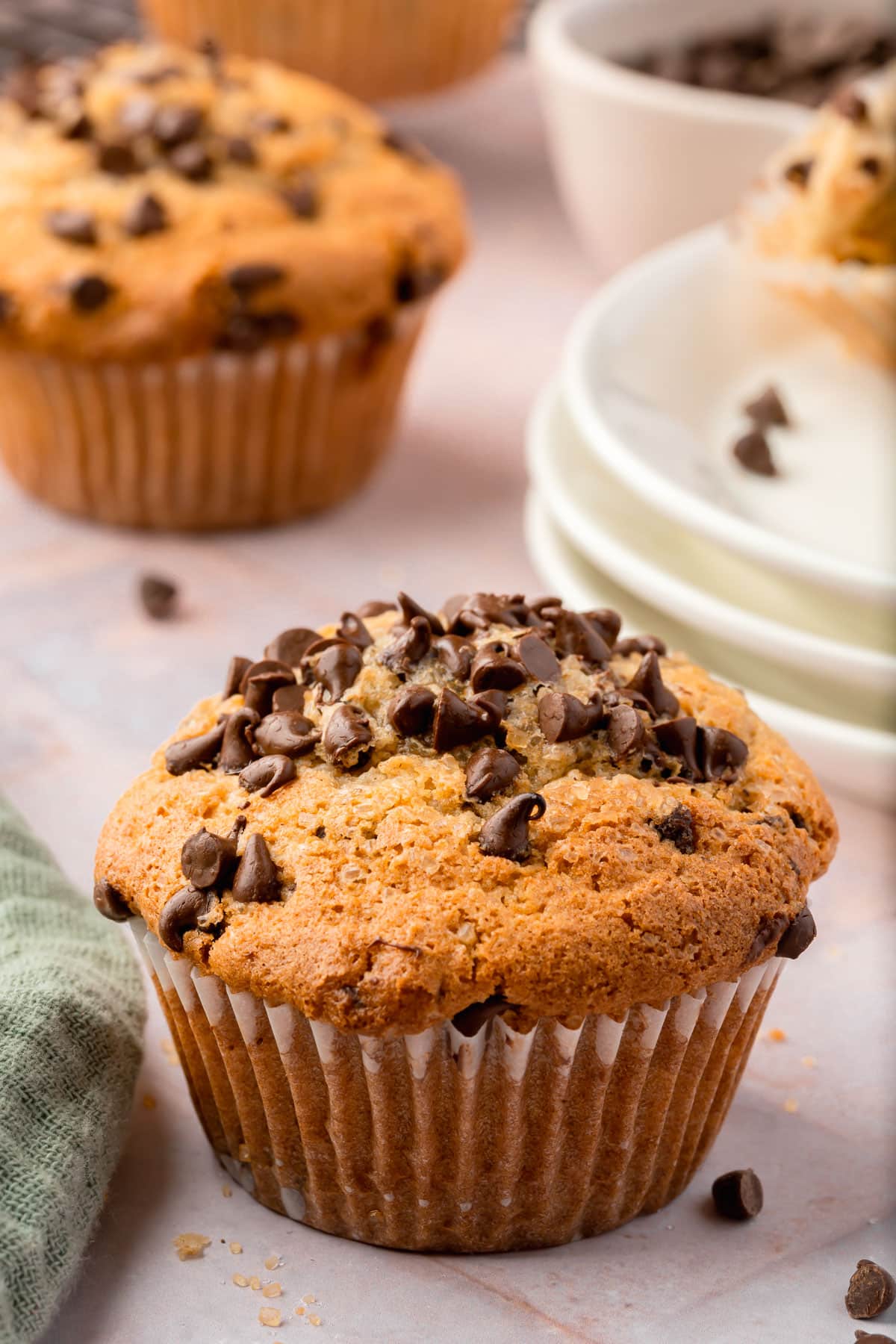 A single gluten-free mini chocolate chip muffin with a stack of dessert plates behind it.
