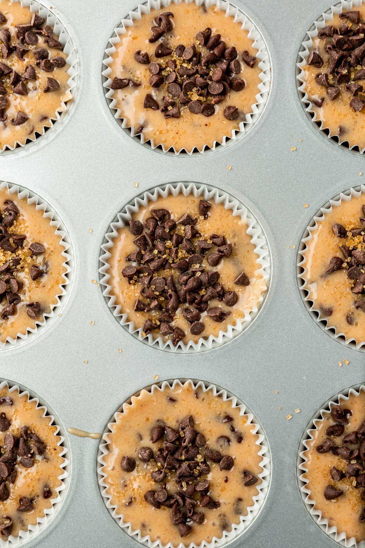 A closeup of muffin liners filled with gluten-free muffin batter topped with mini chocolate chips and turbinado sugar before baking in the oven.