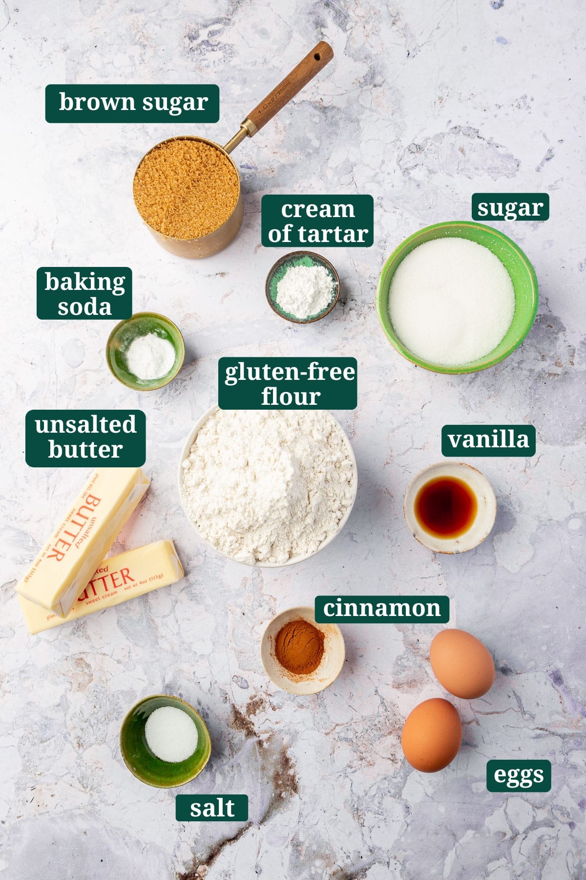 Ingredients in small bowls to make gluten-free snickerdoodles, including brown sugar, granulated sugar, gluten-free flour blend, cream of tartar, butter, eggs, vanilla, cinnamon, baking soda, and salt with text overlays over each ingredient.