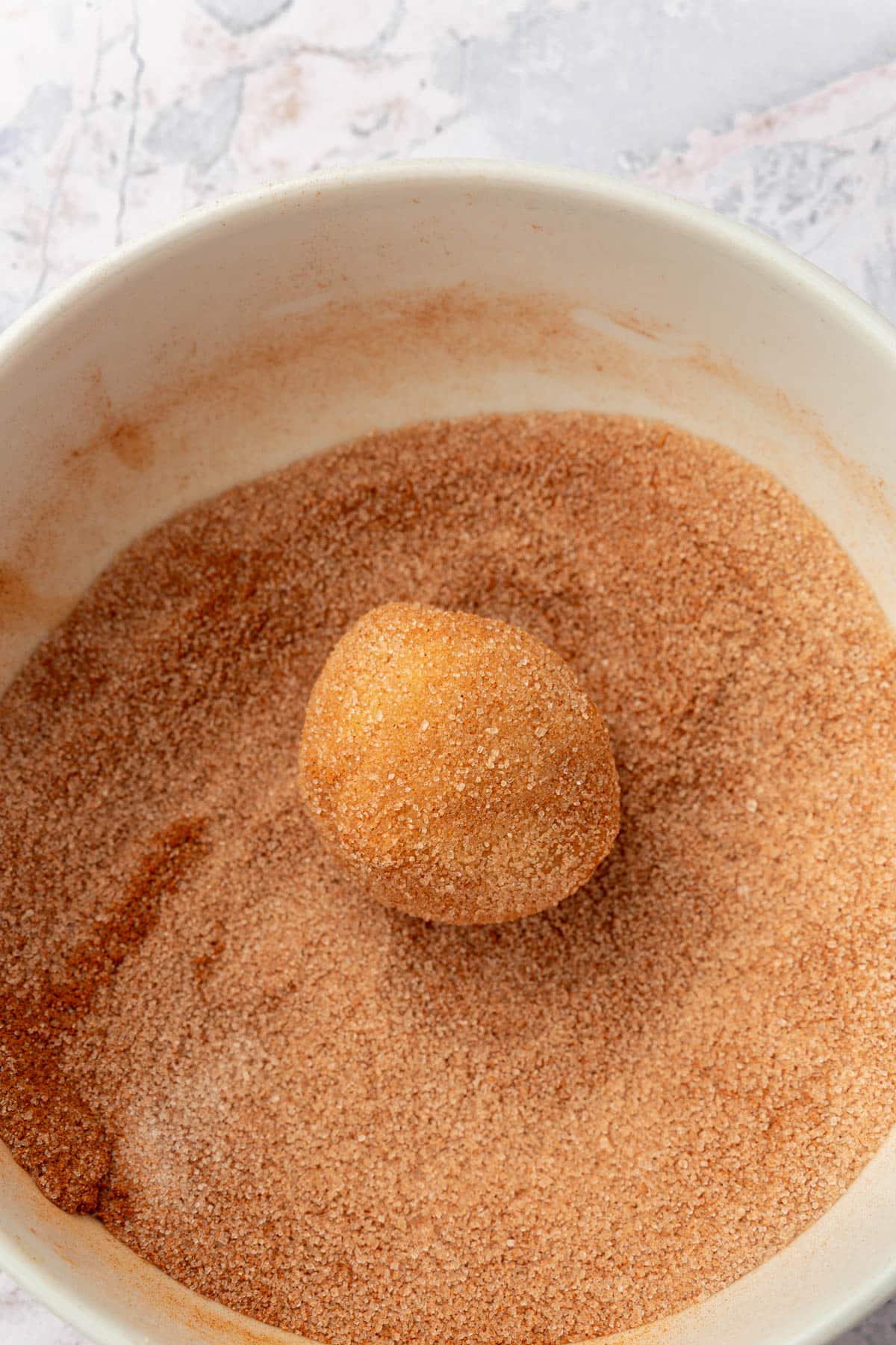 A gluten-free snickerdoodle cookie dough ball rolled in a bowl of cinnamon sugar.