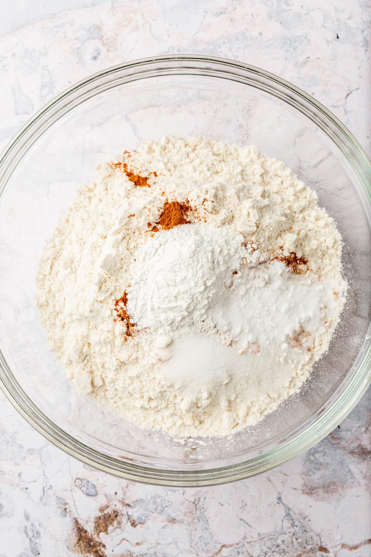 A glass mixing bowl with gluten-free flour, cinnamon, cream of tartar, salt, and baking soda in it before whisking together.