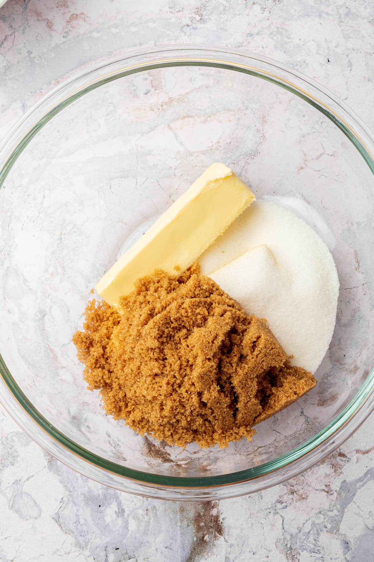 A glass mixing bowl with a stick of butter, granulated sugar, and brown sugar in it before mixing together.