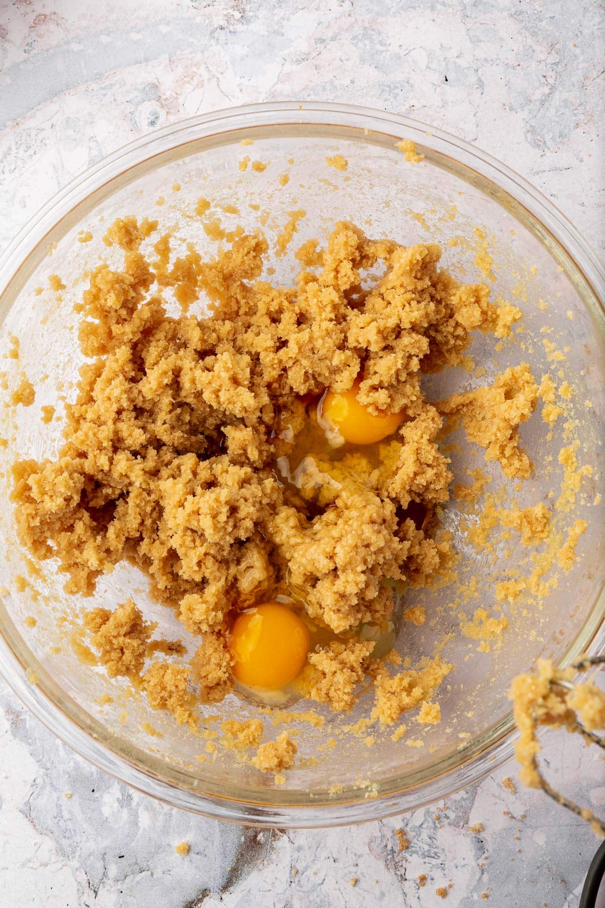 A glass mixing bowl with a creamed butter and brown sugar mixture topped with two raw eggs before mixing together.