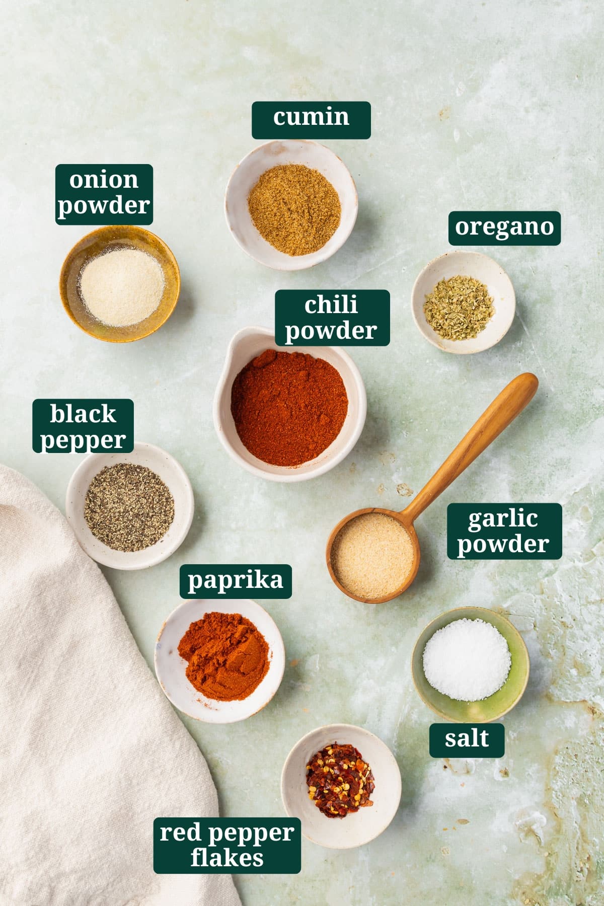 Ingredients in small bowls on a green marble table to make gluten-free taco seasoning, including cumin, onion powder, chili powder, oregano, black pepper, paprika, garlic powder, red pepper flakes and salt with text overlays over each ingredient.