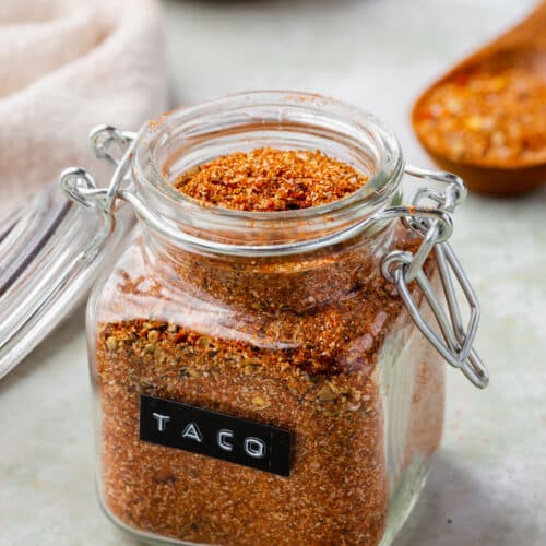 Gluten-free taco seasoning in a glass jar with a black and white label that says "taco" on it with spoons and bowls of jars in the background.