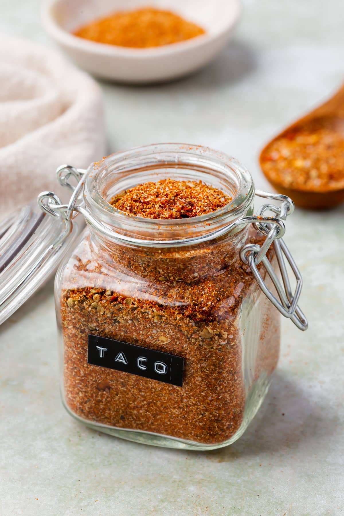Gluten-free taco seasoning in a glass jar with a black and white label that says "taco" on it with spoons and bowls of jars in the background.