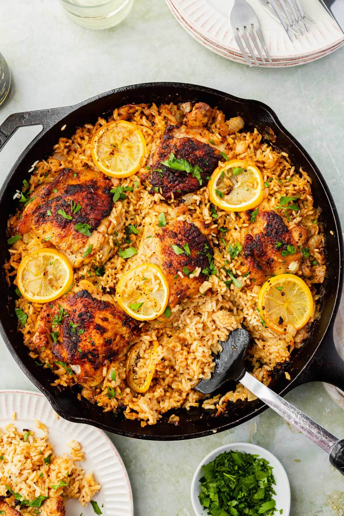 A close up of roasted chicken thighs on top of a bed of seasoned white rice and topped with charred lemon slices and fresh parsley in a cast iron skillet.