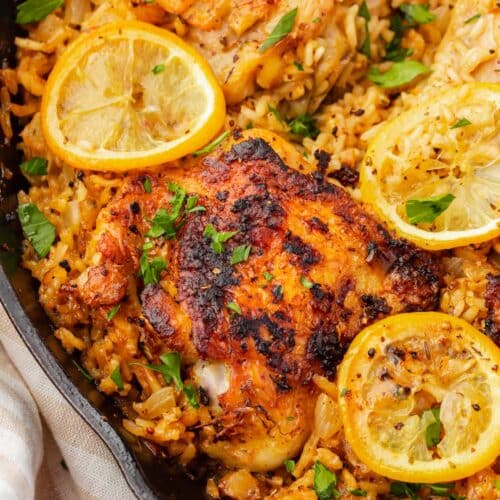 A close up of roasted chicken thighs on top of a bed of seasoned white rice and topped with charred lemon slices and fresh parsley in a cast iron skillet.