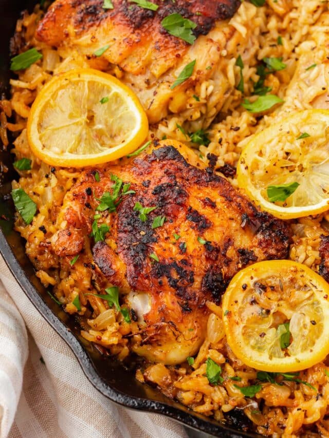 A close up of roasted chicken thighs on top of a bed of seasoned white rice and topped with charred lemon slices and fresh parsley in a cast iron skillet.