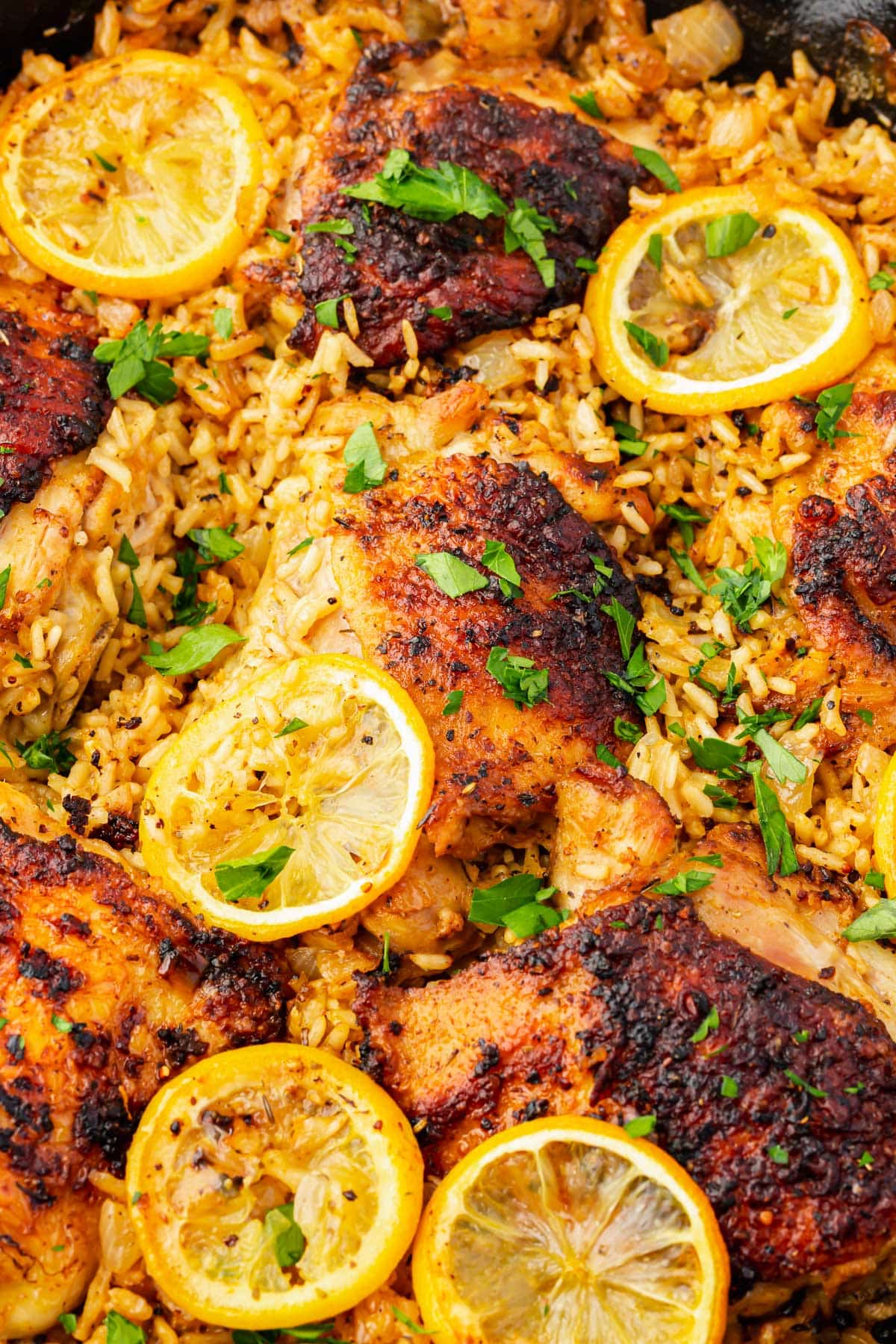 A close up of roasted chicken thighs on top of a bed of seasoned white rice and topped with charred lemon slices and fresh parsley in a cast iron skillet.