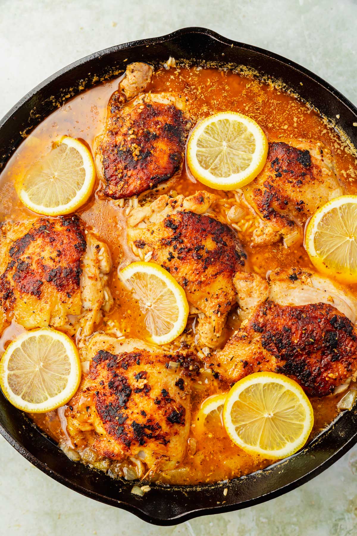 A cast iron skillet with charred chicken thighs, rice, fresh lemon slices and chicken broth in it before baking in the oven.
