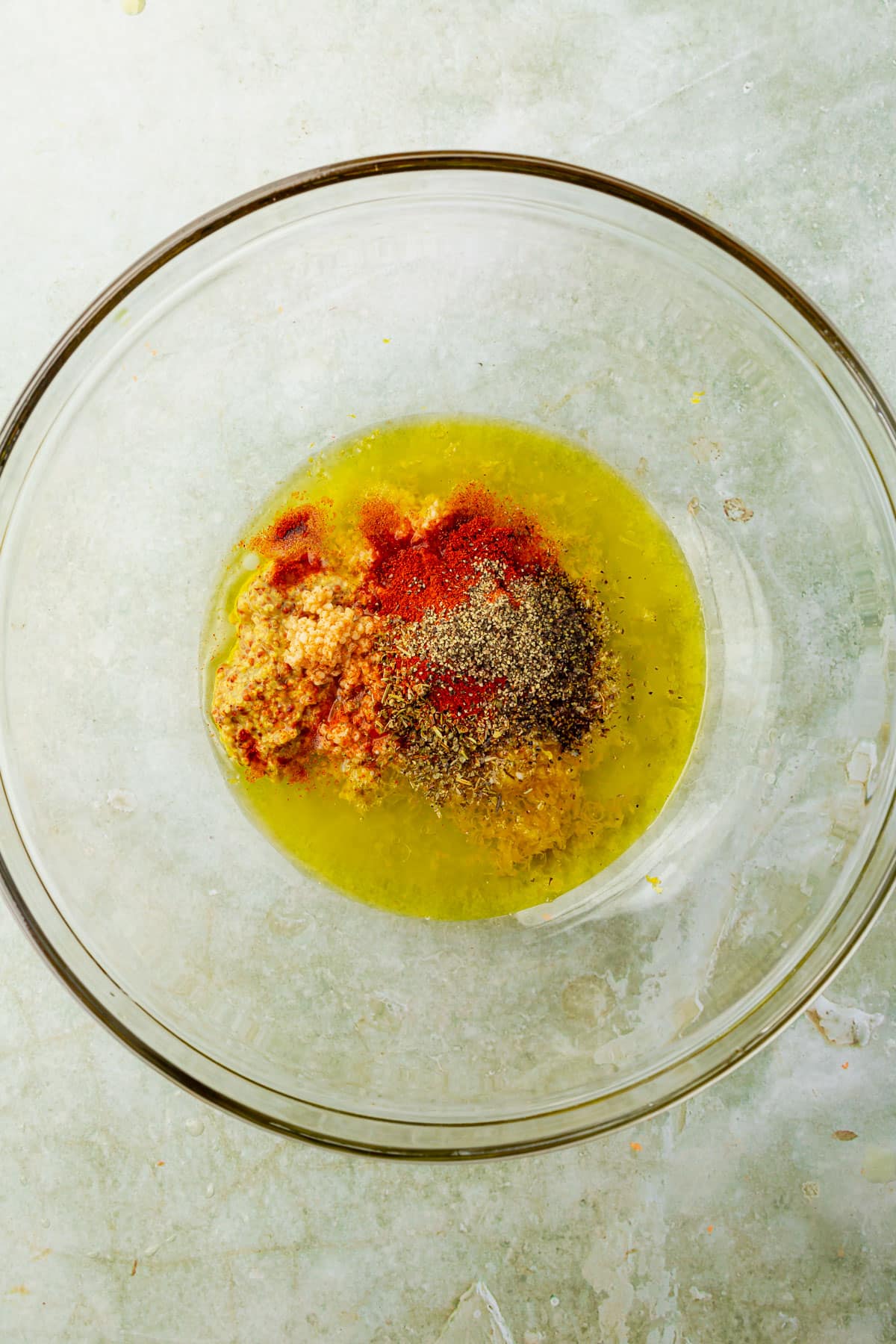 A glass mixing bowl with lemon juice, olive oil, paprika, garlic, Italian seasoning blend, lemon peel, and black pepper in it before mixing together.