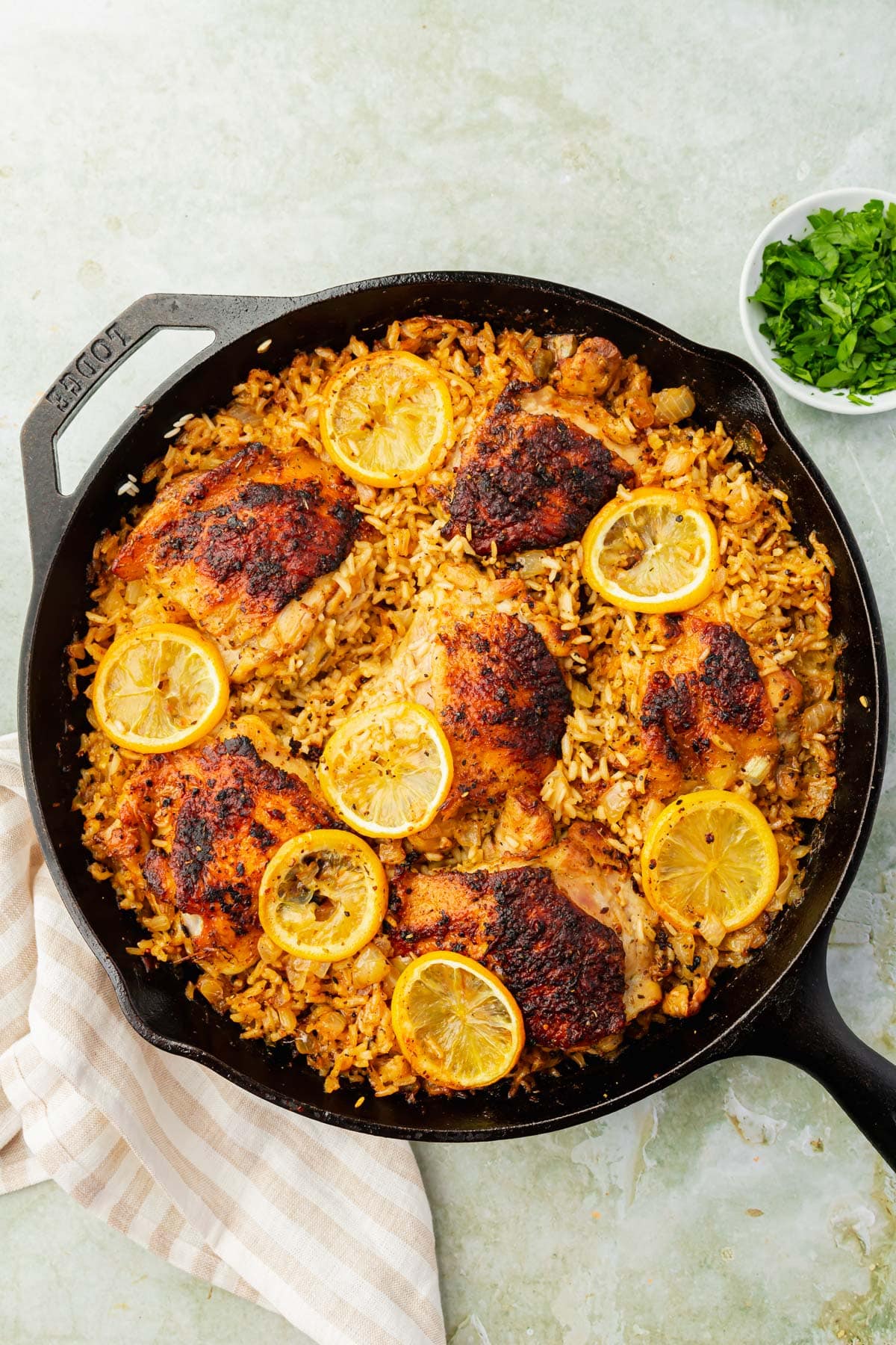 A cast iron skillet with rice, roasted chicken thighs, lemon slices and a small bowl of fresh chopped parsley to the side.