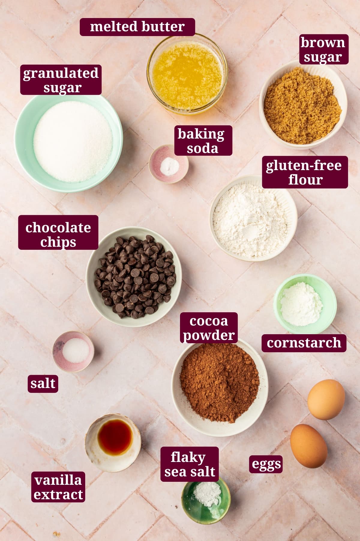 An overhead view of ingredients in small bowls to make gluten-free brownies, including granulated sugar, melted butter, brown sugar, baking soda, gluten-free flour, chocolate chips, cocoa powder, cornstarch, eggs, and vanilla extract, with text overlays over each ingredient.