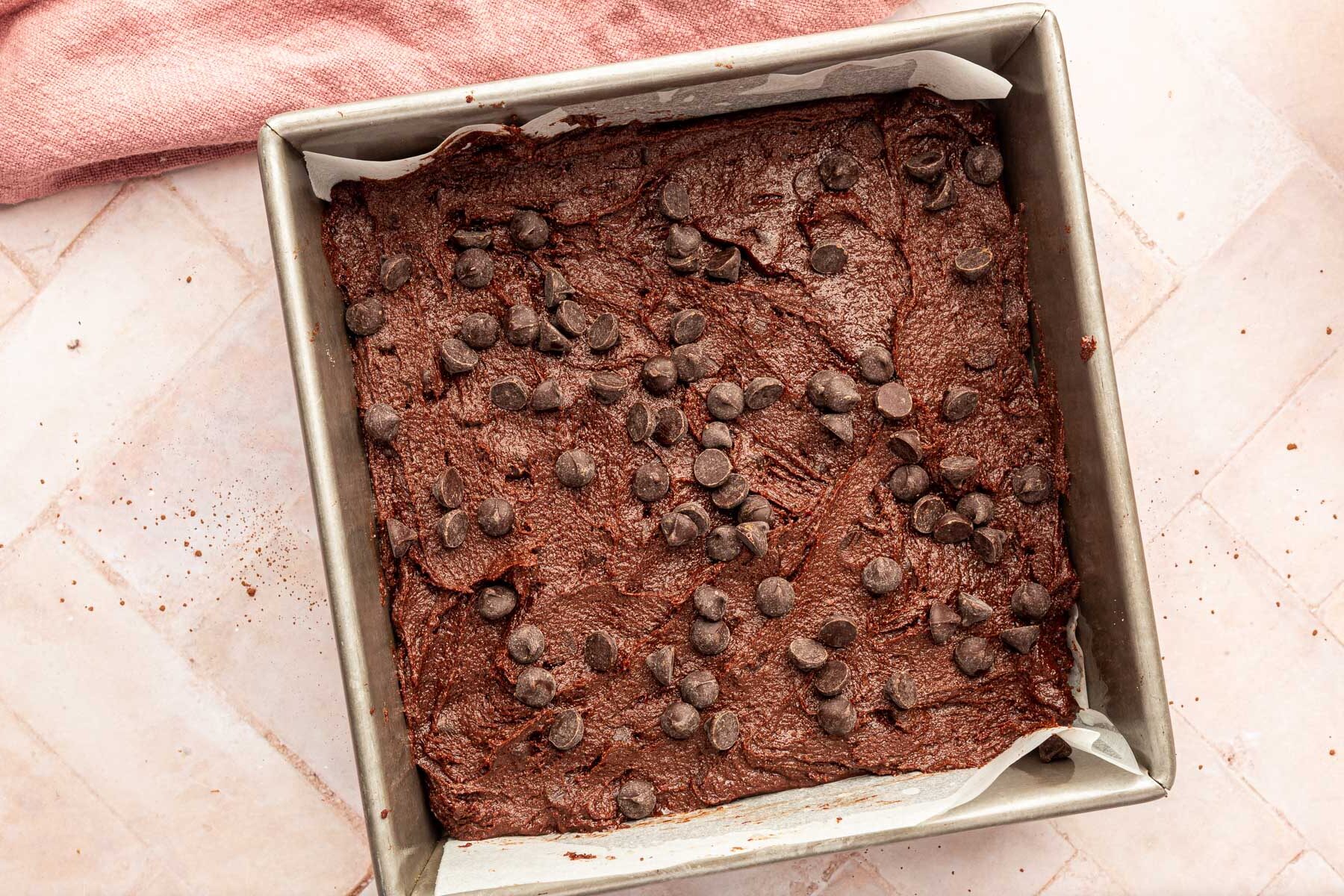 A square baking pan filled with brownie batter and topped with semi-sweet chocolate chips before baking in the oven.