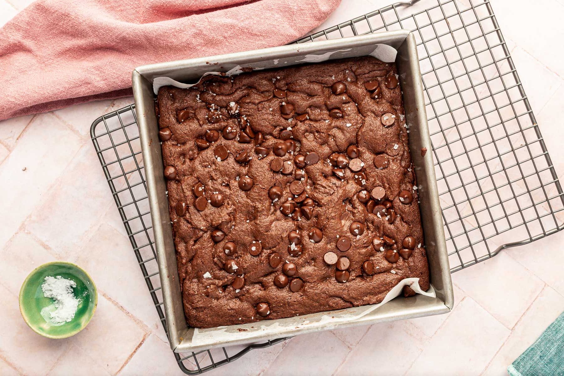 A square baking pan filled with baked gluten-free brownies with a sprinkle of flaky sea salt on top of a pink background.