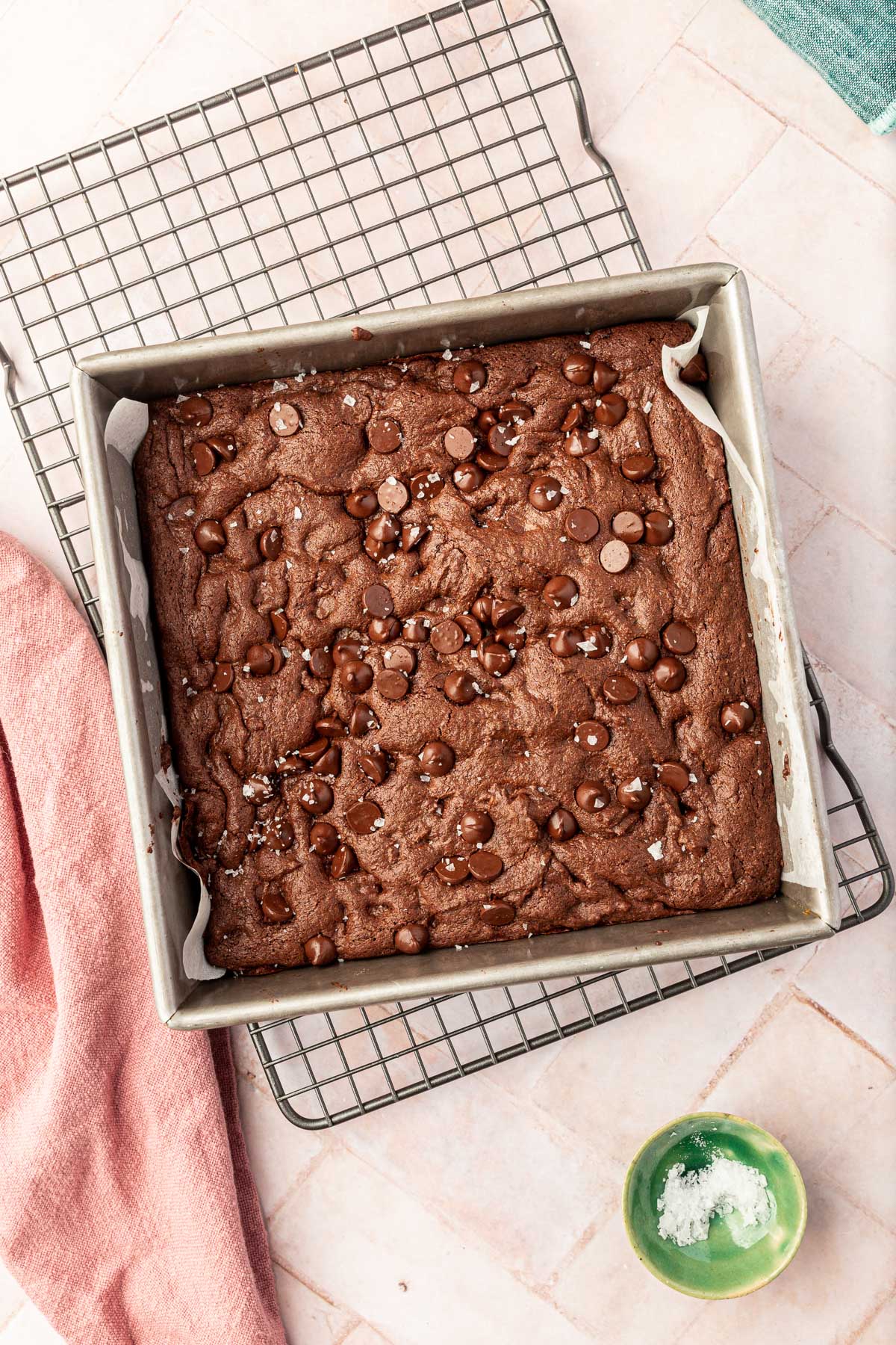 A square baking pan filled with baked gluten-free brownies with a sprinkle of flaky sea salt on top of a pink background.