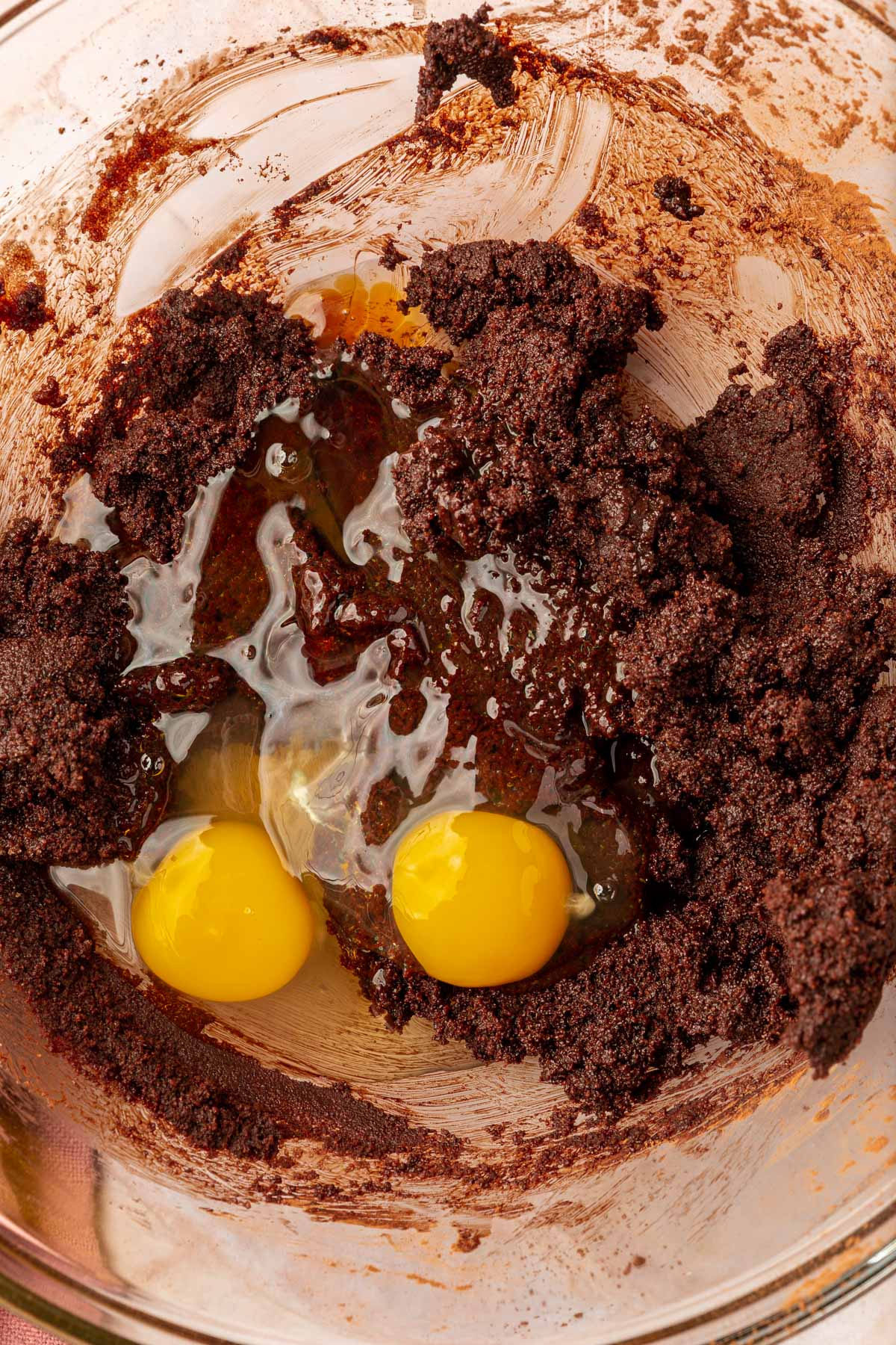 A glass mixing bowl with a chocolate brownie mixed topped with two eggs before mixing together.