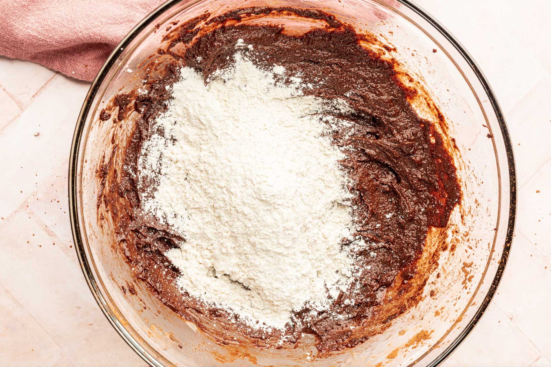 A glass mixing bowl with brownie batter topped with a pile of gluten-free flour before mixing together.