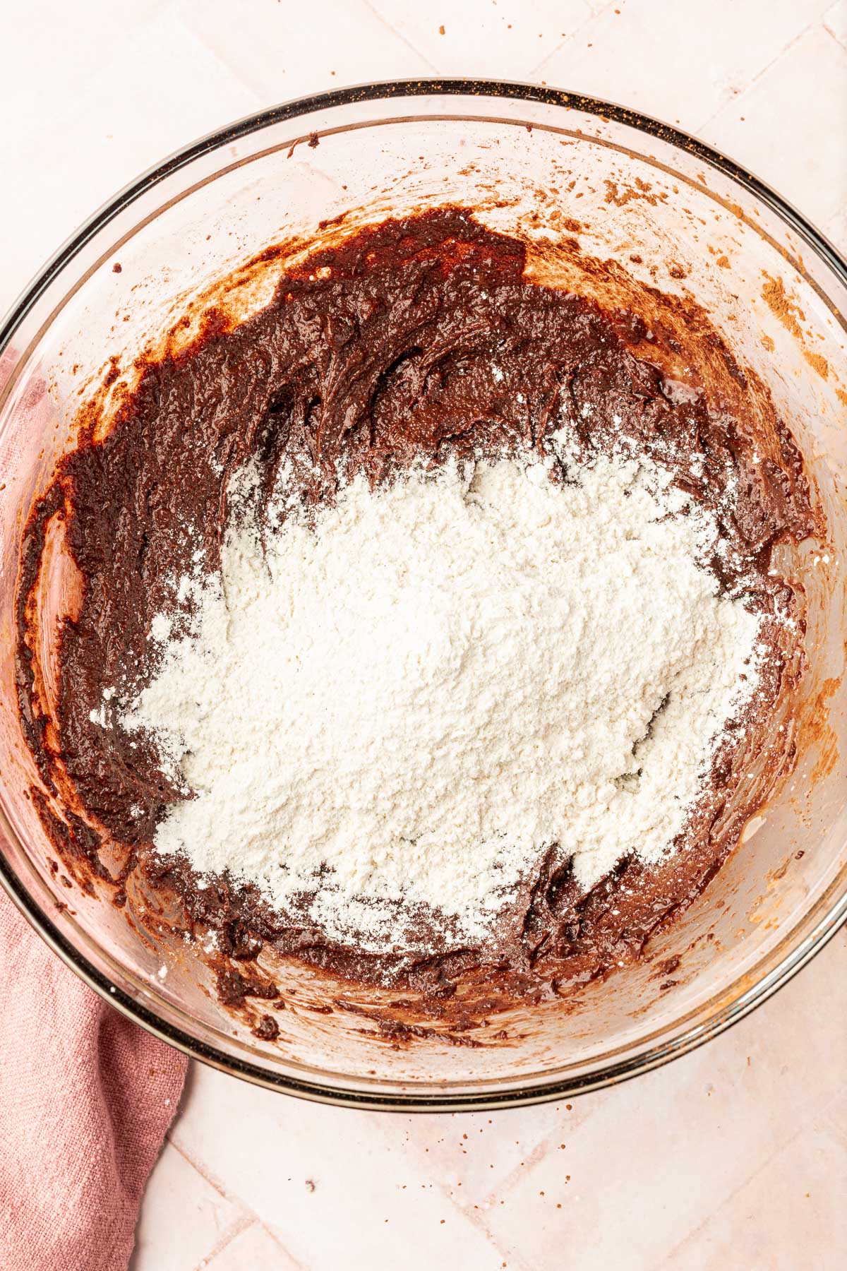 A glass mixing bowl with brownie batter topped with a pile of gluten-free flour before mixing together.