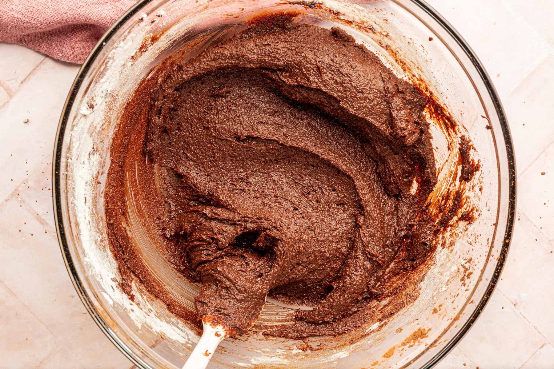 A glass mixing bowl filled with gluten-free brownie batter with a pink spatula.