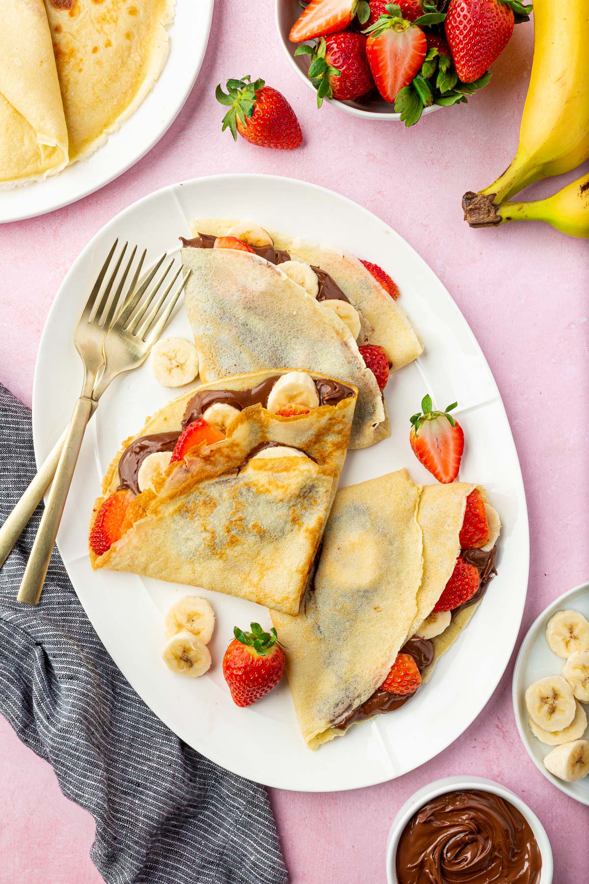 A platter with three gluten-free crepes filled with nutella, banana slices, and sliced strawberries with a few forks.