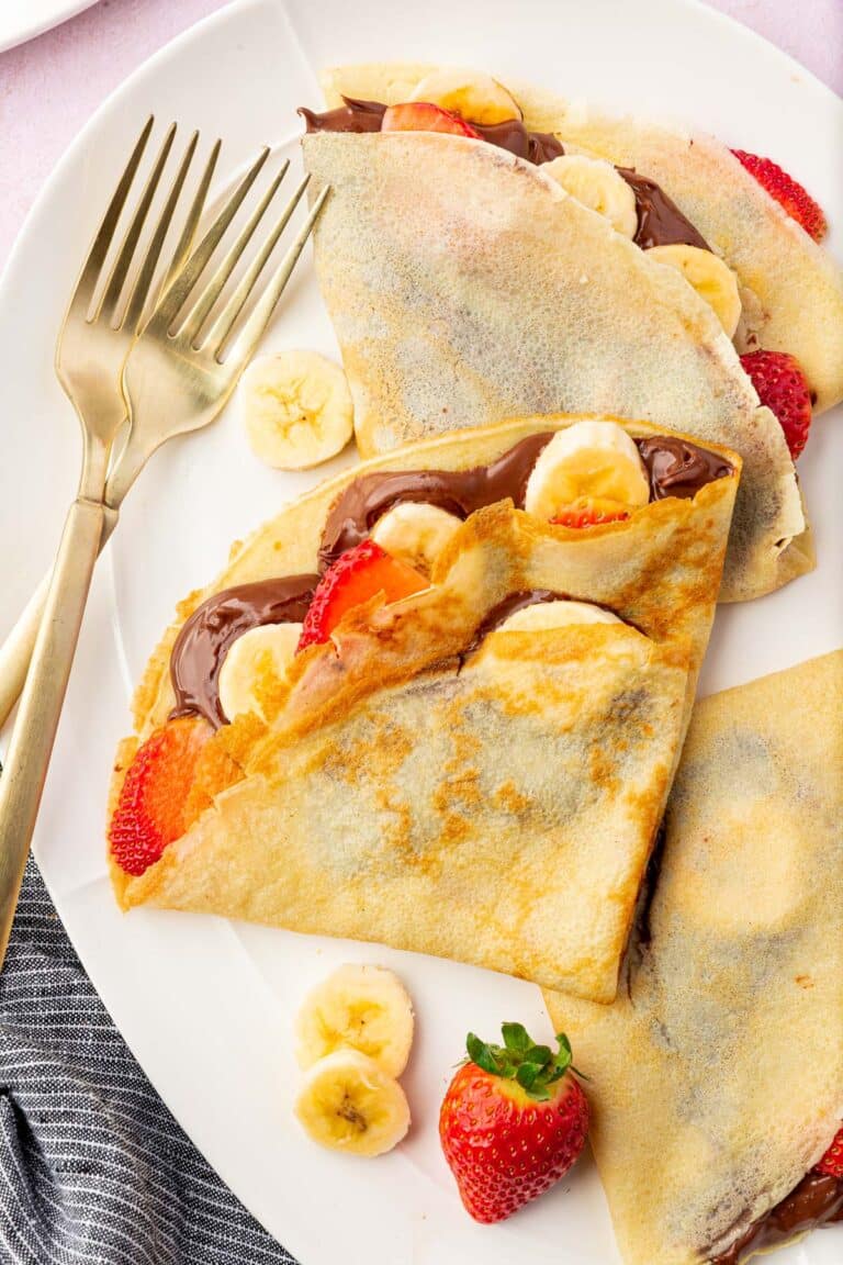 A platter with three gluten-free crepes filled with nutella, banana slices, and sliced strawberries with a few forks.