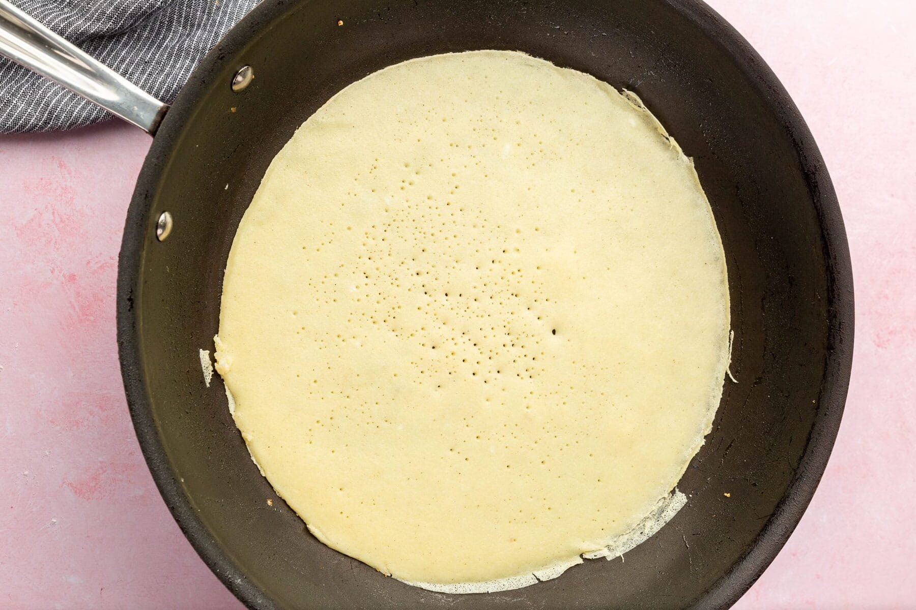 A thin crepe cooking in a black nonstick skillet, with small bubbles on the surface, resting on a light pink countertop next to a gray-striped cloth.