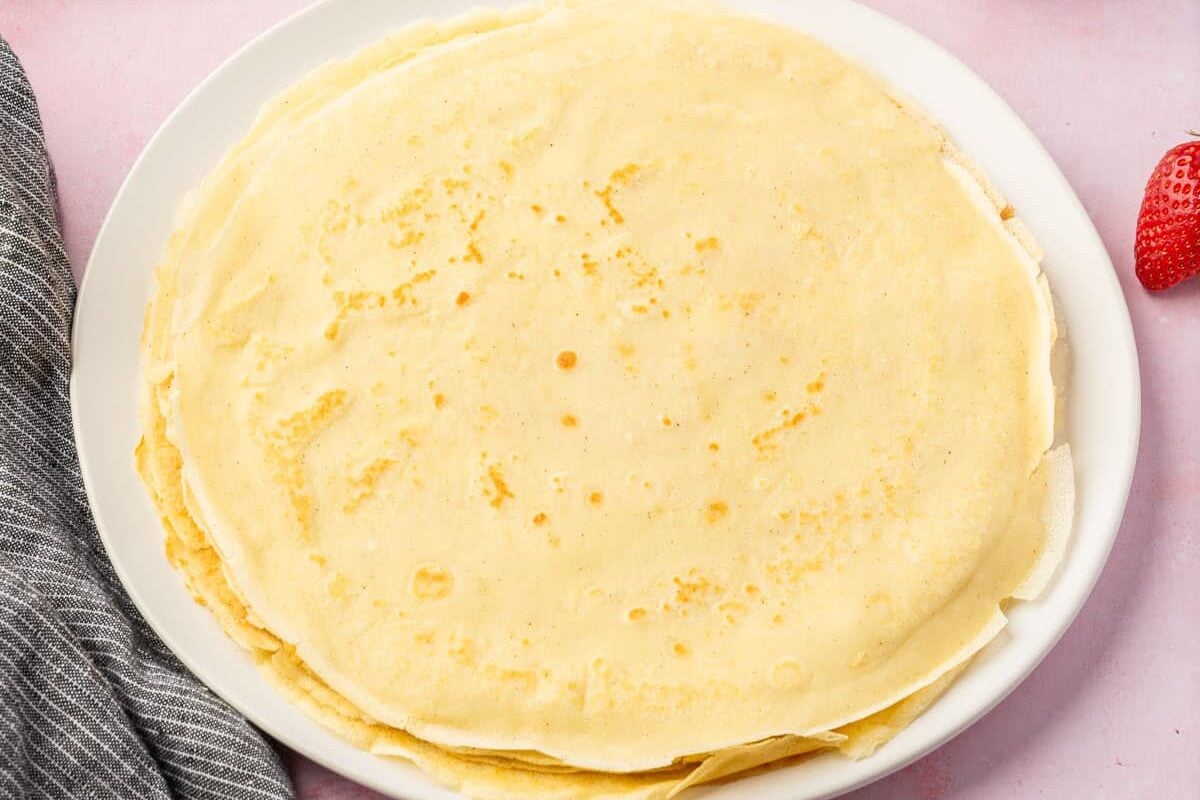A stack of thin, golden crepes sits on a white plate, with a gray-striped cloth on the side and a single strawberry partially visible in the corner on a light pink surface.