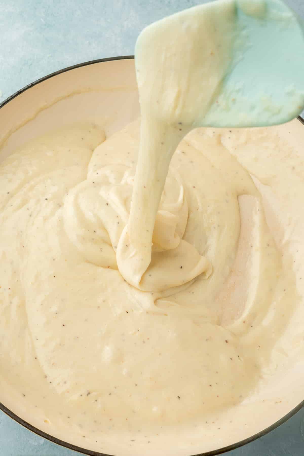 An overhead view of a white cheese sauce in a white skillet being drizzled from overhead by a blue spatula.
