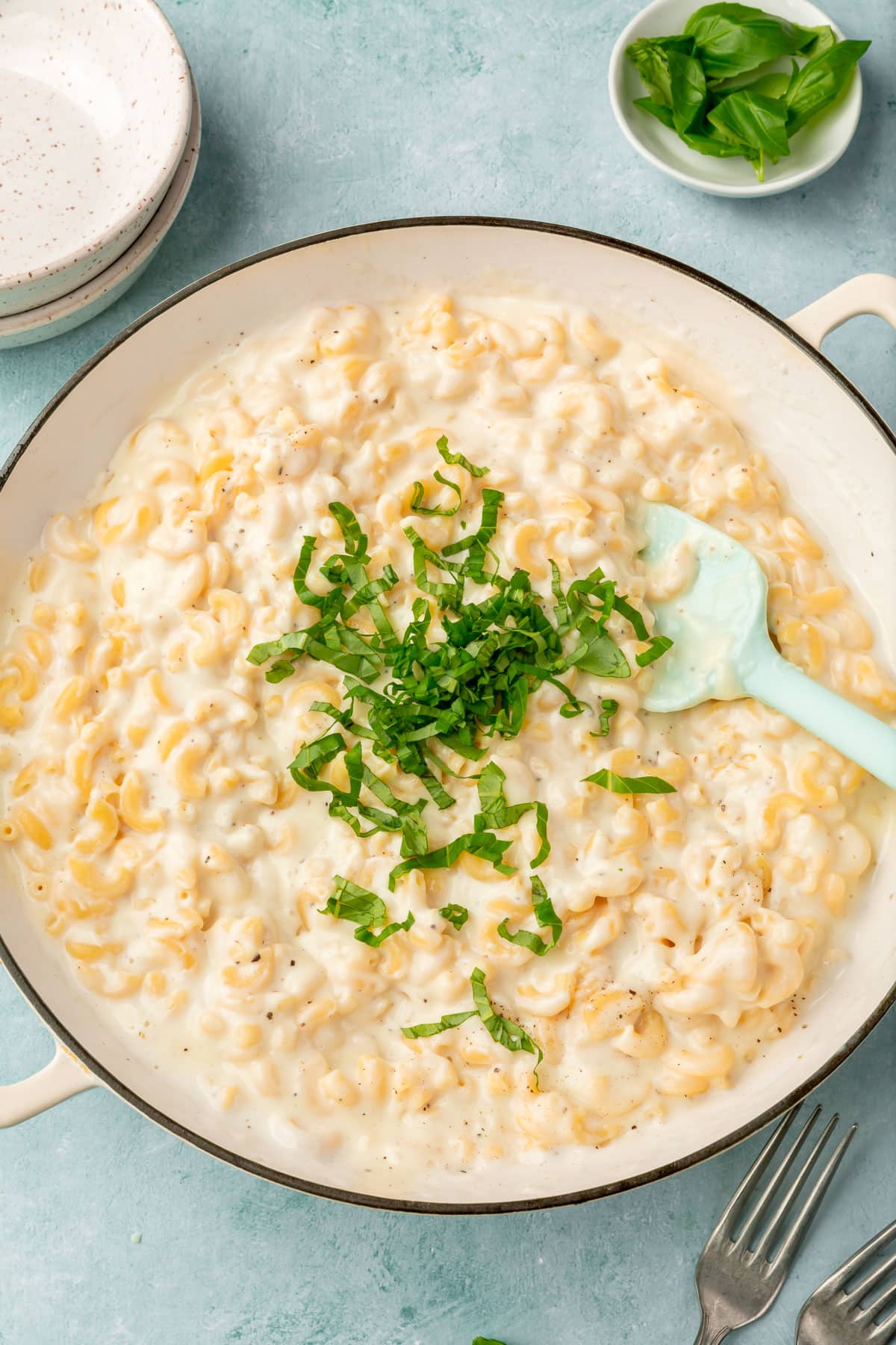 A white dutch oven with mozzarella mac and cheese with a pile of thinly sliced basil in it with a blue silicone spatula to mix it together.