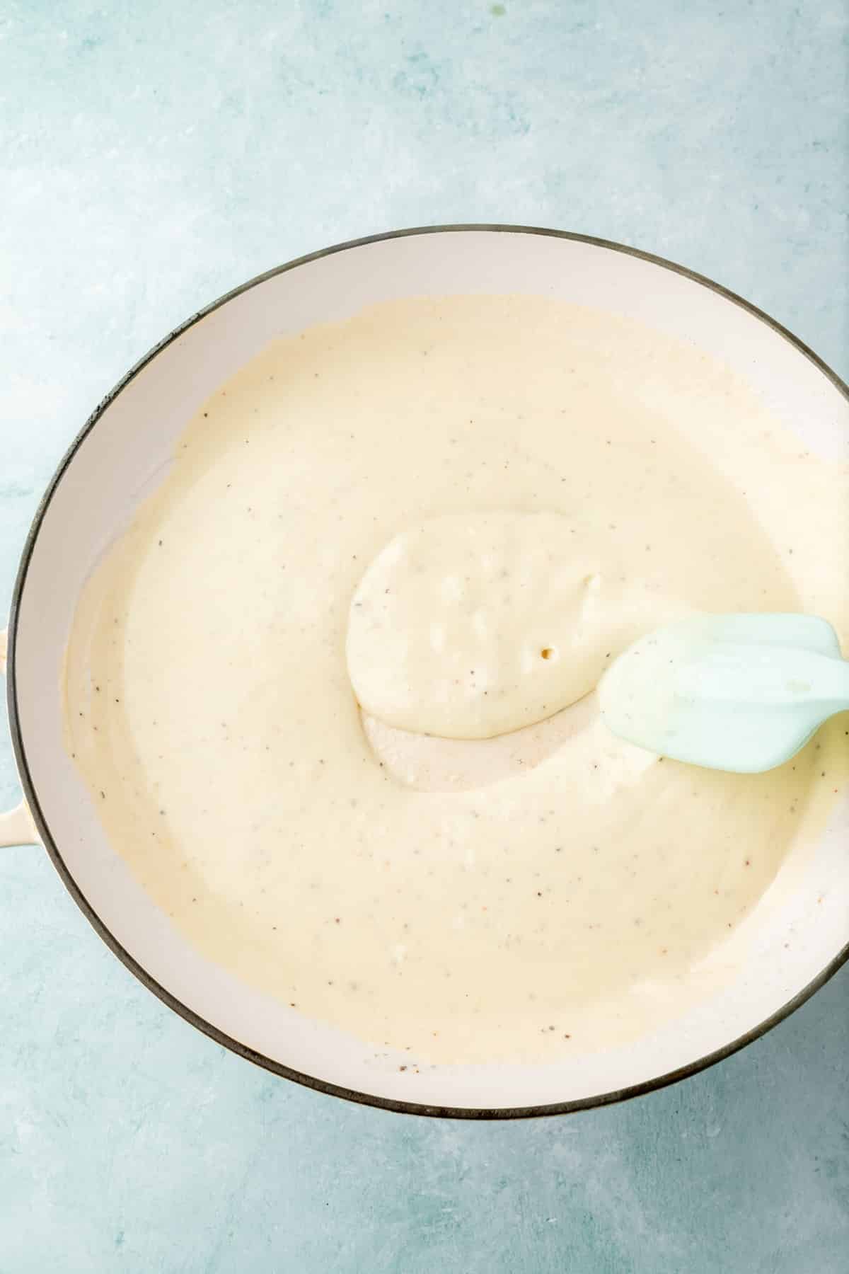 A white cream sauce being stirred together with a blue spatula in a white skillet.