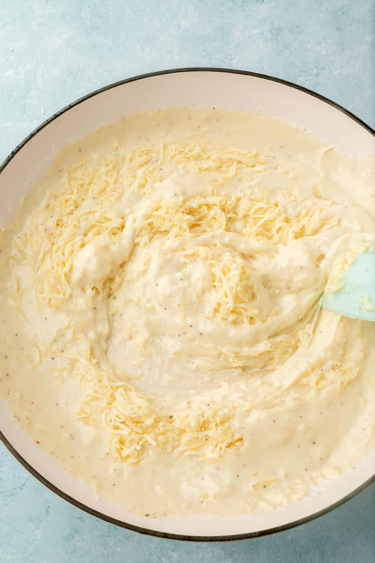 An overhead view of shredded mozzarrella cheese being stirred into a white cream sauce with a blue spatula.