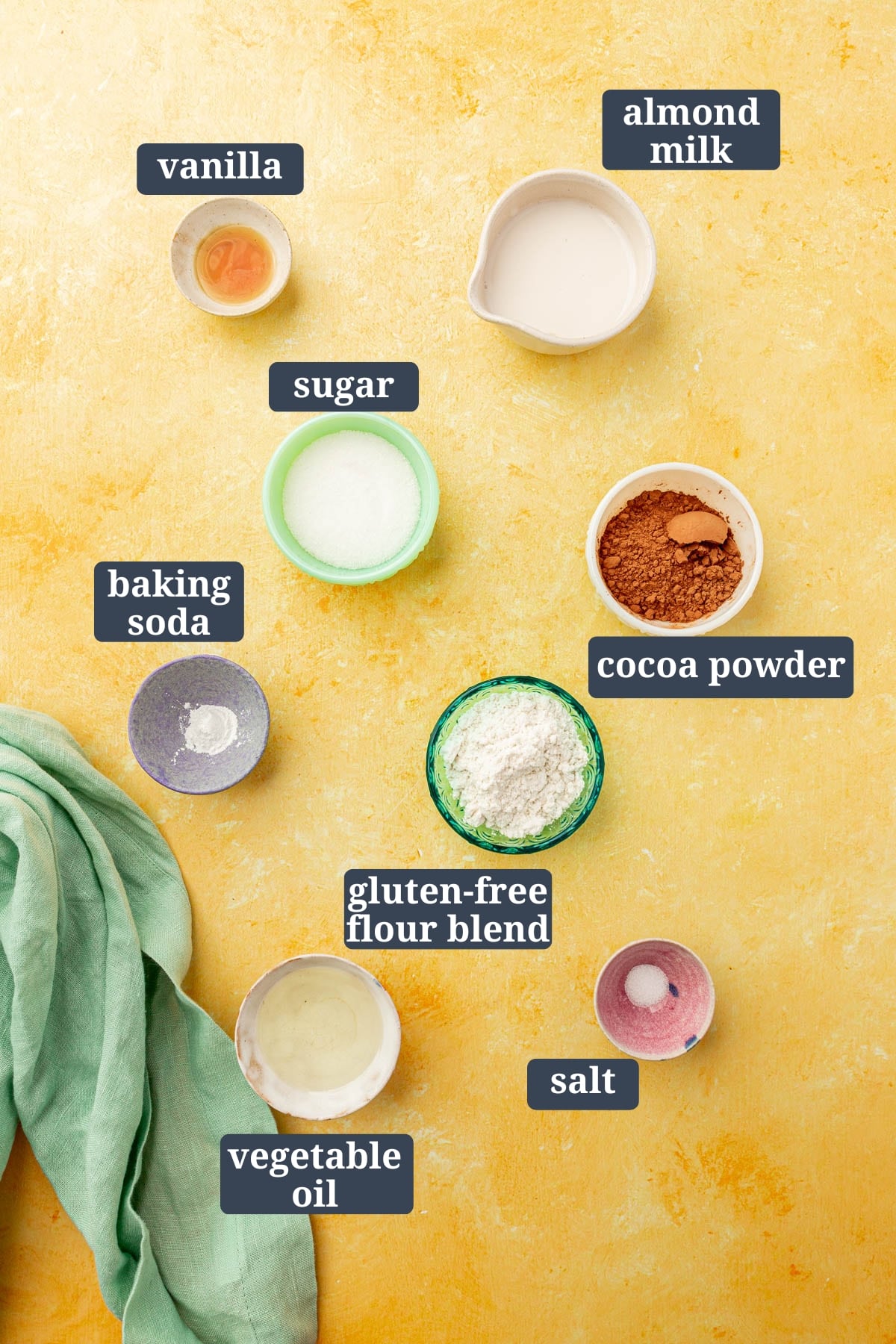 Ingredients in small colorful bowls to make a gluten-free chocolate mug cake, including cocoa powder, oil, sugar, gluten-free flour, baking soda, vanilla, almond milk with text overlays over each ingredient.