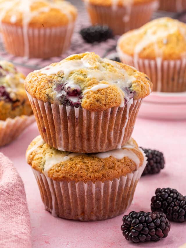 A stack of two gluten-free blackberry muffins with lemon glaze with additional muffins and fresh blackberries in the background.