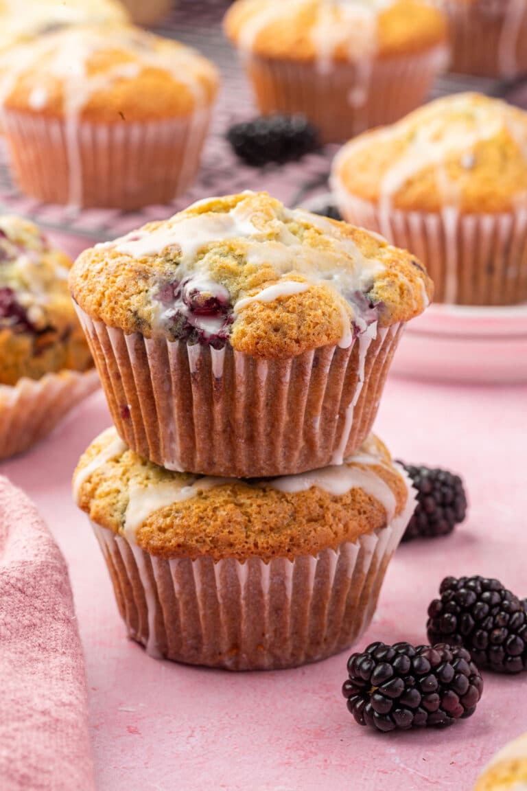 A stack of two gluten-free blackberry muffins with lemon glaze with additional muffins and fresh blackberries in the background.