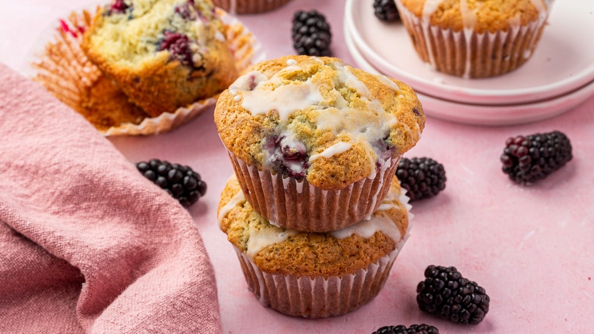 Two lemon blackberry muffins stacked on top of each other with fresh blackberries and additional muffins in the background.
