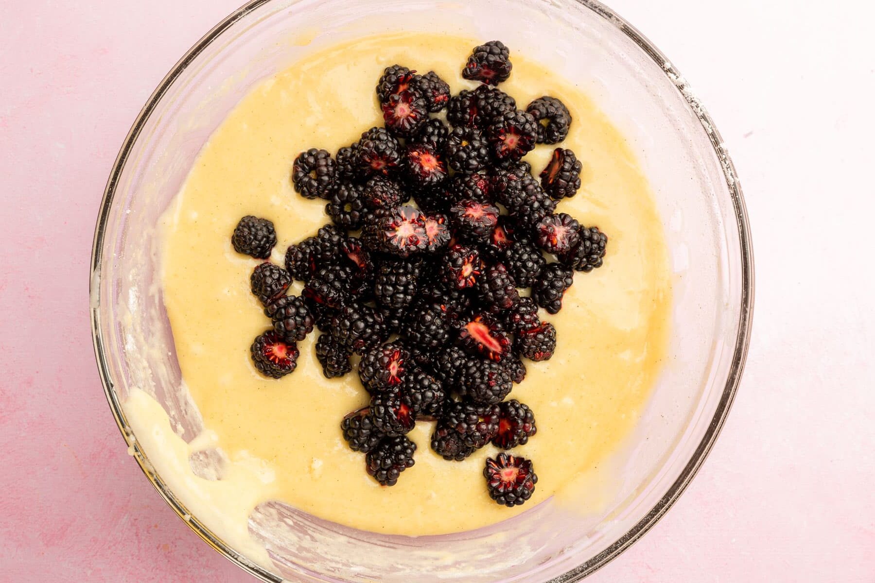 A glass mixing bowl filled with gluten-free muffin batter topped with chopped blackberries before mixing together.